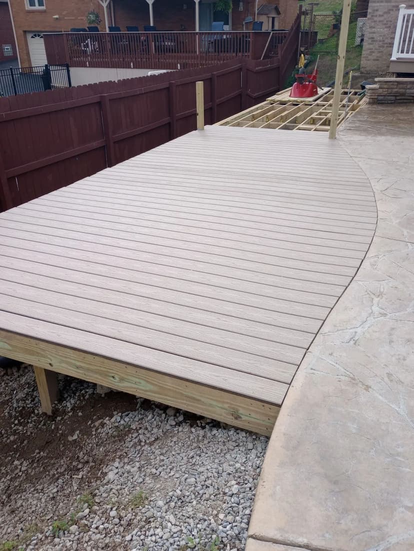 Curved wooden deck under construction in a backyard with a concrete patio nearby.