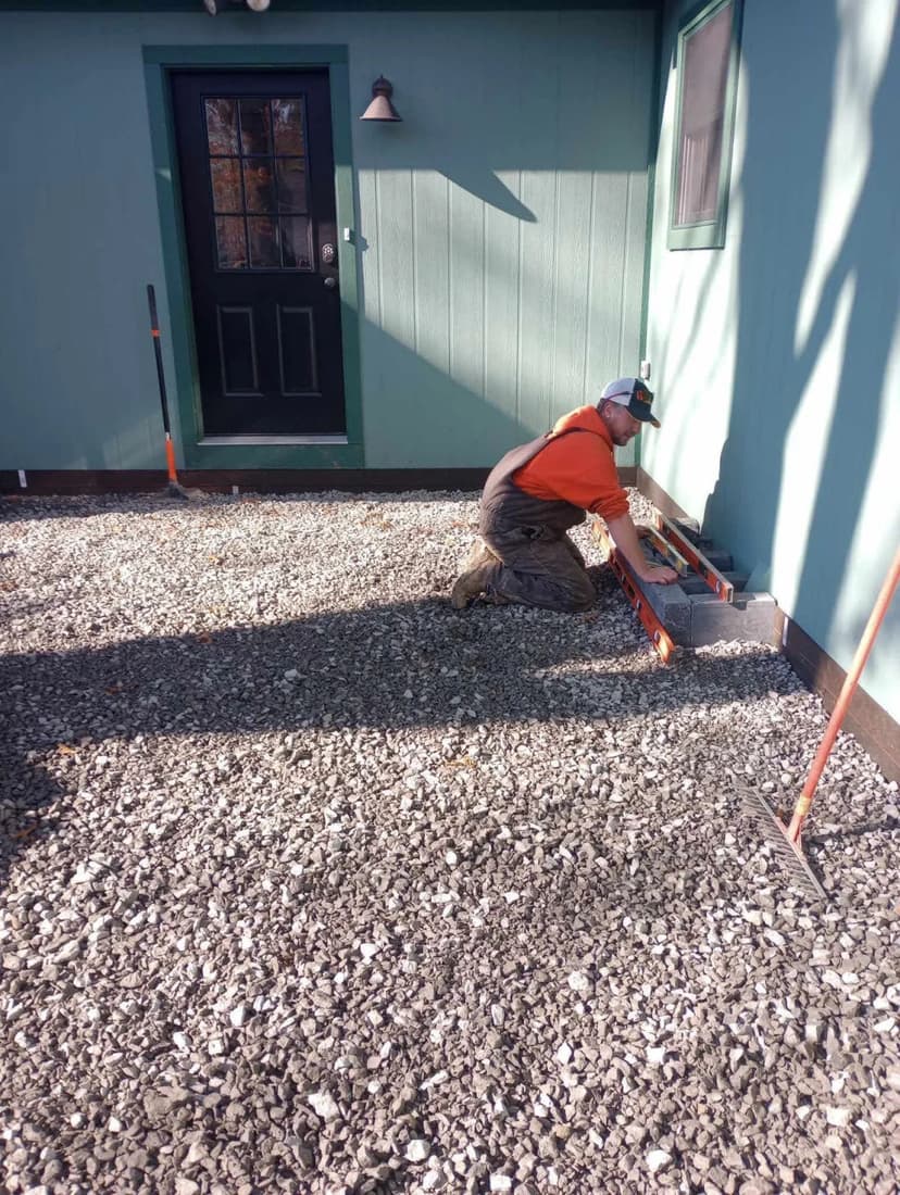 Person working outside on gravel near a green building, measuring and leveling ground.